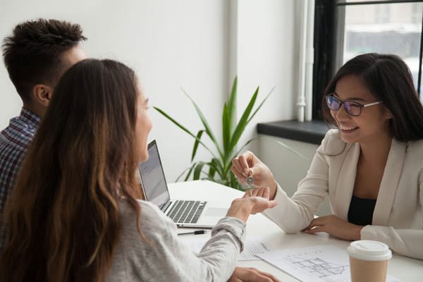 Three colleagues in a bright office sit around a table; a woman with glasses hands keys to a client while a laptop and papers lie nearby.