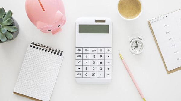 Desk with a white calculator surrounded by a piggy bank, notebook, clock, coffee, calendar, pencil, and succulent plant.