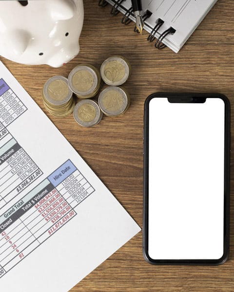 Piggy bank with coins beside financial documents and a blank smartphone on a wooden desk.